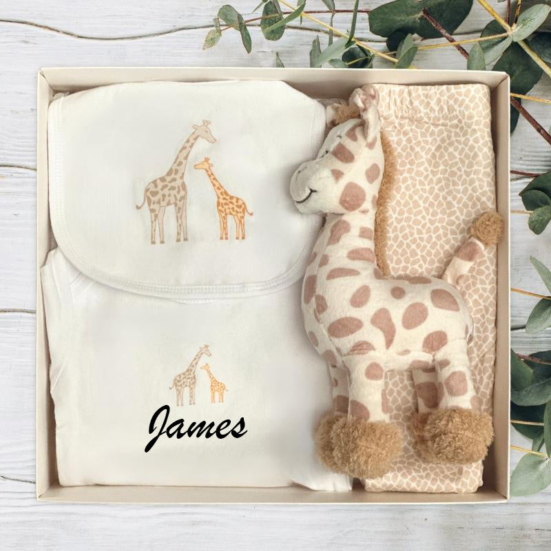 Gift set with plush giraffe toy and baby clothing in a gift box on a wooden surface.