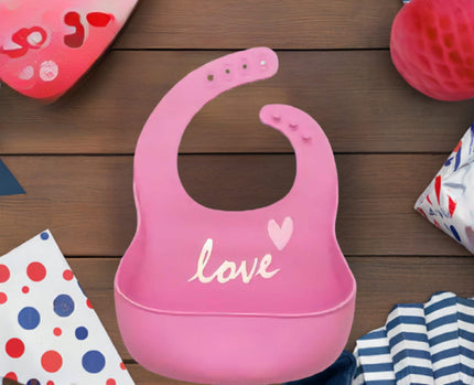 Pink baby bib with 'love' and a heart on a wooden surface with colorful items.