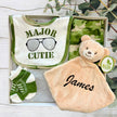 Baby gift set with bib, major cutie nanu clothing, and toy in a box on a wooden surface with greenery.