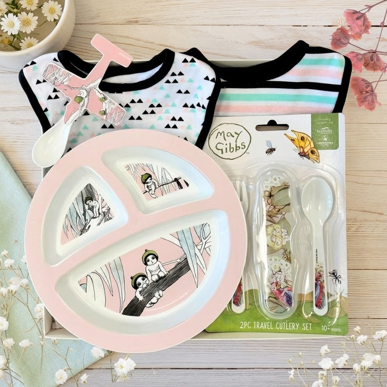 May Gibbs themed baby gift set with Gumnut Babies pink plate, cutlery, and bibs on a wooden surface.