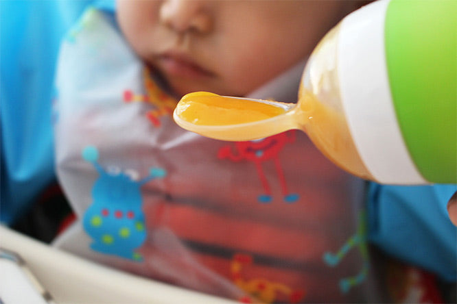 Child using a green and white spoon with orange food, wearing a colorful bib.