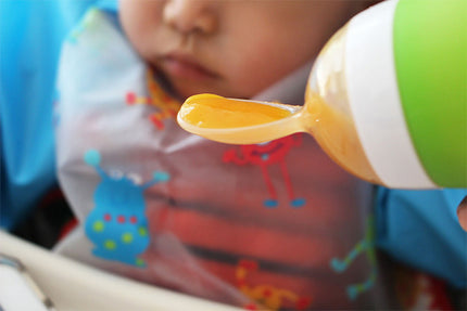 Child using a green and white spoon with orange food, wearing a colorful bib.