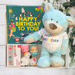 Blue teddy bear with 'Happy Birthday' shirt and children's book in a gift box