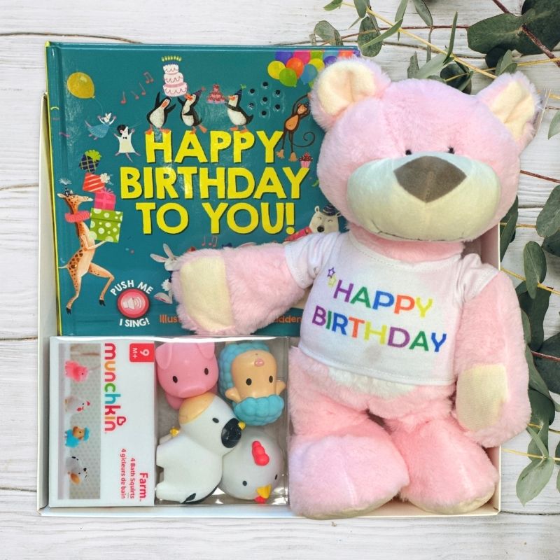 Pink teddy bear with 'Happy Birthday' shirt, children's book, and toy in a gift box on a light wooden surface.