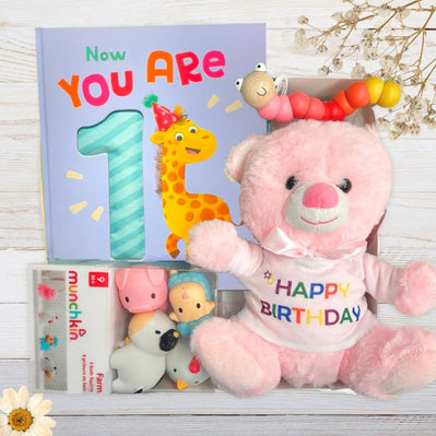 Pink teddy bear with 'Happy Birthday' shirt, children's book, and toy in a gift box on a light wooden surface.