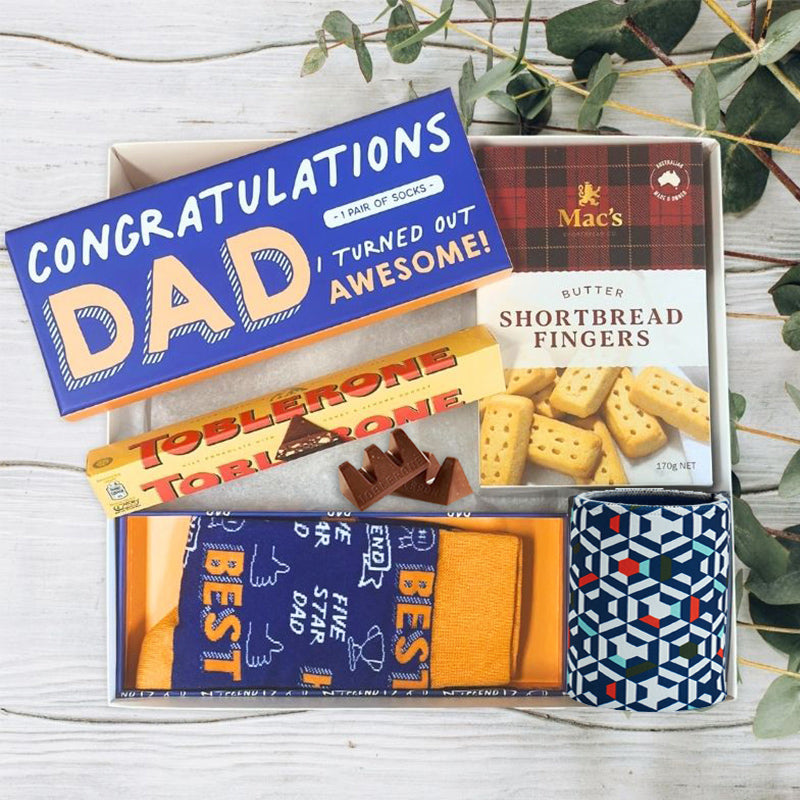 Collection of dad gifts including a box of shortbread fingers, Toblerone chocolate, and a drink cooler, gift box by 'The Baby Gift Company'.