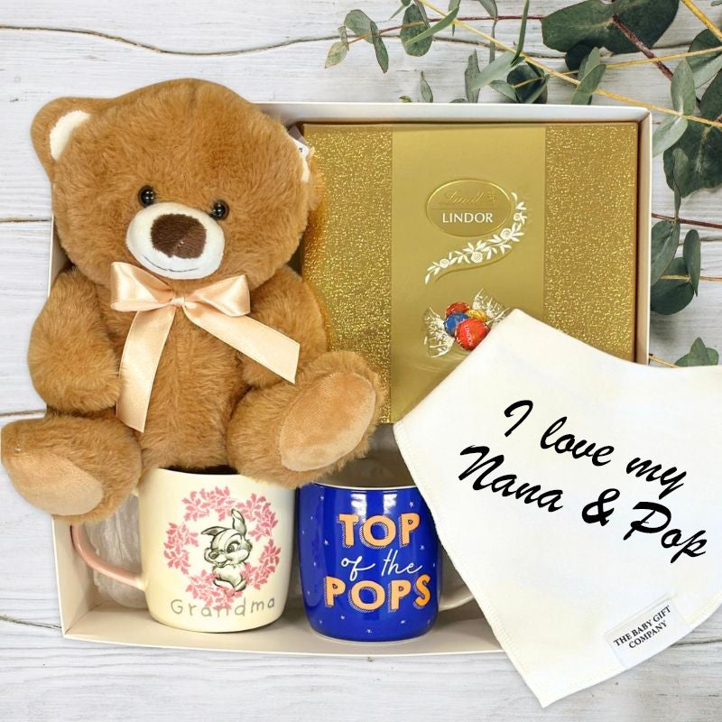 Teddy bear, mugs, Lindor chocolate box, and bib in gift box on a wooden surface with greenery.