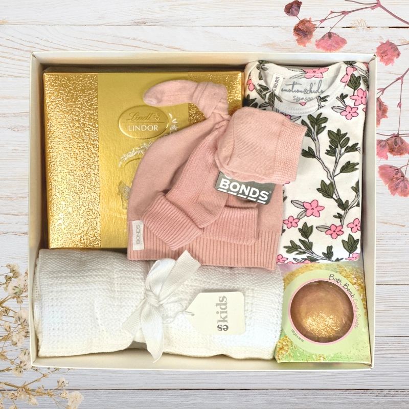 Gift box containing baby items including a pink bonds hat, white blanket, and Lindor chocolate box.
