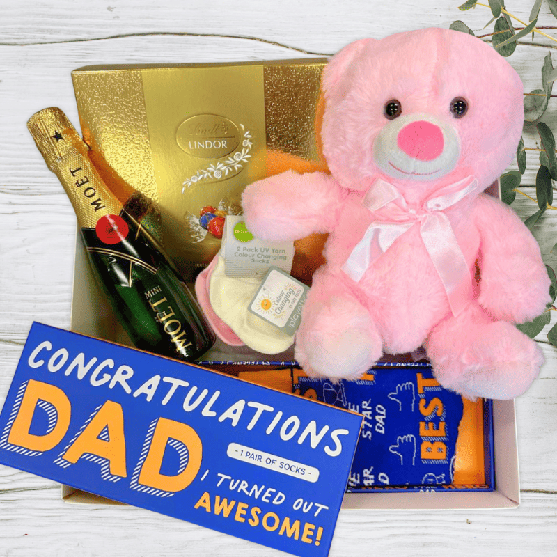 Gift set with pink teddy bear, Moet champagne, Lindor chocolates, and 'Congratulations Dad' socks on a white background