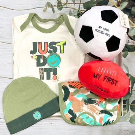 Nike Baby clothing and toys including a onesie, hat, soccer ball, and football on a wooden surface with greenery.