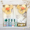 Baby gift set with duck-shaped rattle, bottles, and Ferrero Rocher on a wooden surface.