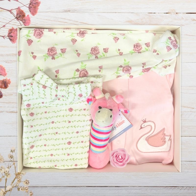 Baby gift box with floral clothing, pink swan cardigan, and giraffe toy in a box on a wooden surface.