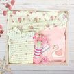 Baby gift box with floral clothing, pink swan cardigan, and giraffe toy in a box on a wooden surface.