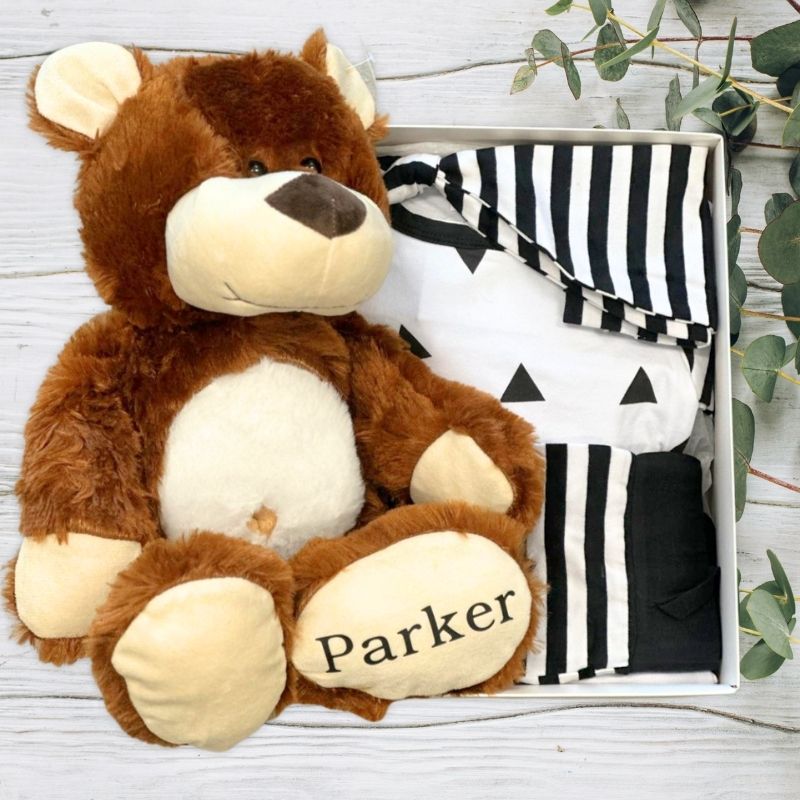 Brown teddy bear with 'Parker' on its paw, sitting next to black and white striped babywear on a wooden surface.