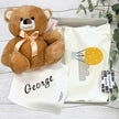 Brown teddy bear with a bow and a white baby bib with 'George' on it, placed in gift box on a wooden surface with greenery.