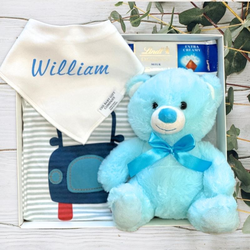 Blue teddy bear with a bow, baby blanket, and Lindt chocolate in a gift box on a wooden surface.