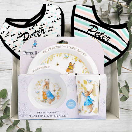 Peter Rabbit-themed mealtime dinner set with plates, bowls, and bibs on a wooden surface.