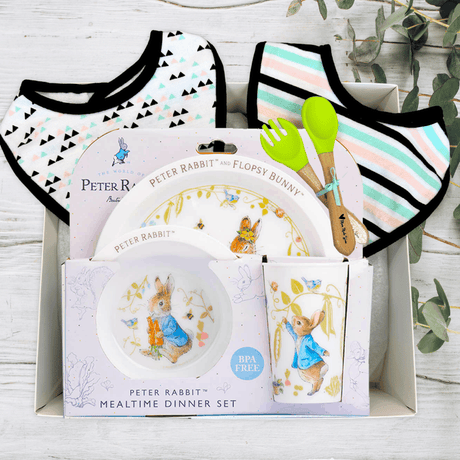 Peter Rabbit-themed mealtime dinner set with plates, bowls, and bibs on a wooden surface.