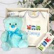Baby gift set with blue teddy bear, baby clothes, and train-themed item on a light wooden surface.