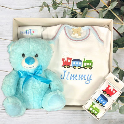 Baby gift set with blue teddy bear, baby clothes, and train-themed item on a light wooden surface.