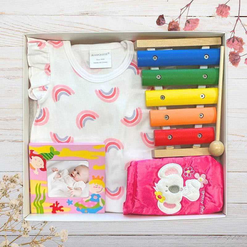 Baby girl clothing set with a colourful xylophone in a baby gift box on a wooden surface.