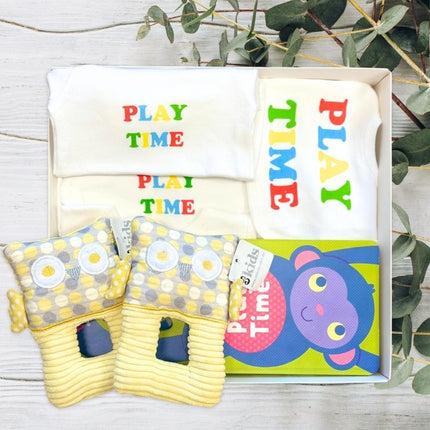 Baby playtime clothes including rompers, bibs, and toys in gift box on a wooden surface with greenery.