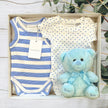 Baby gift set with clothes and a teddy bear in a box on a wooden surface.