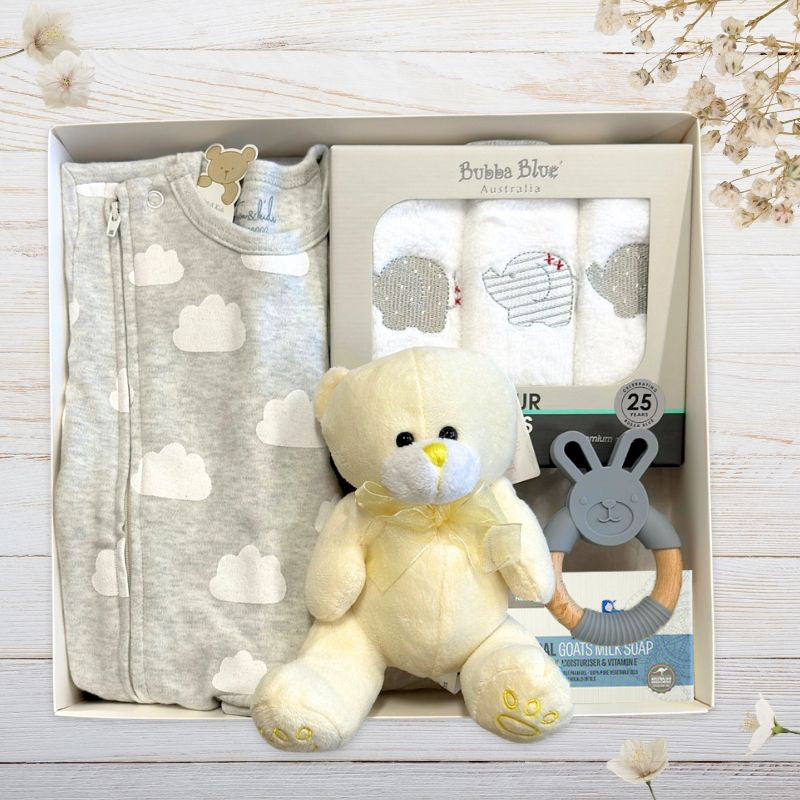 Baby gift set with grey sleeper, teddy bear, and teether in gift box on a light wooden surface.