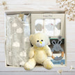 Baby gift set with grey sleeper, teddy bear, and teether in gift box on a light wooden surface.