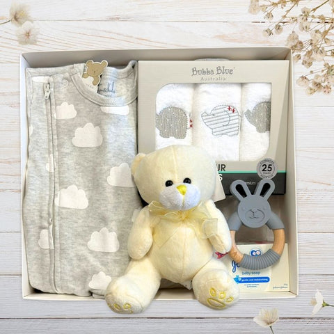 Baby gift set with grey sleeper, teddy bear, and teether in gift box on a light wooden surface.
