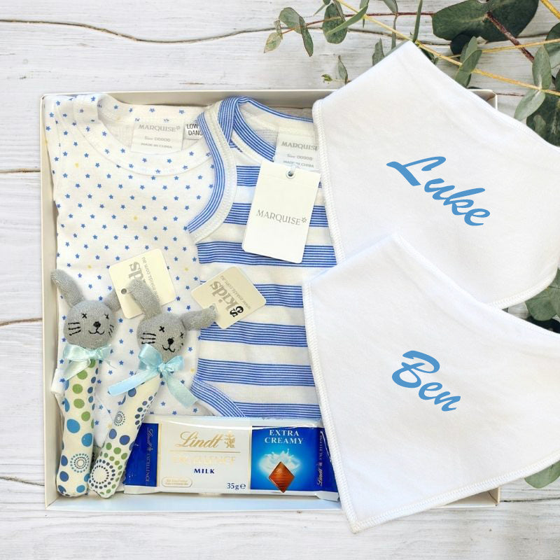 Baby gift set with clothes, toys, and personalized baby bibs for twin boys on a wooden surface.