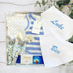 Baby gift set with clothes, toys, and personalized baby bibs for twin boys on a wooden surface.