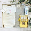Baby gift set with clothes, a toy, and a bottle in a box on a wooden surface with greenery.