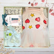 Baby outfit with dress, shoes, and items in a box on a wooden surface.