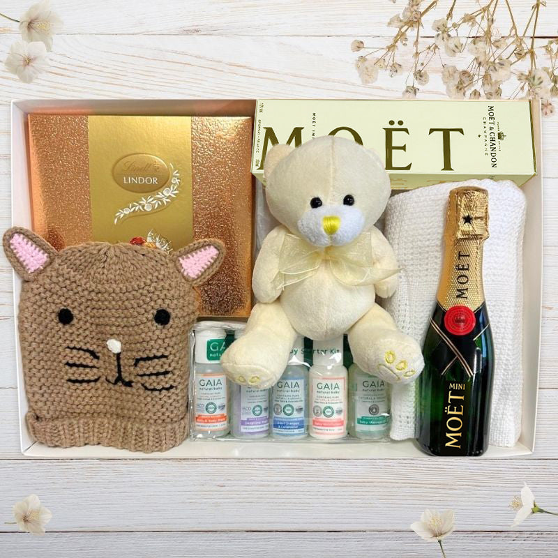 Baby Gift set with teddy bear, cat-shaped knitted hat, Moét champagne, and gaia skincare on a wooden surface.