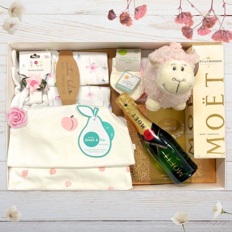 Gift box with baby items, Moet champagne, and a lamb plush toy on a wooden surface.