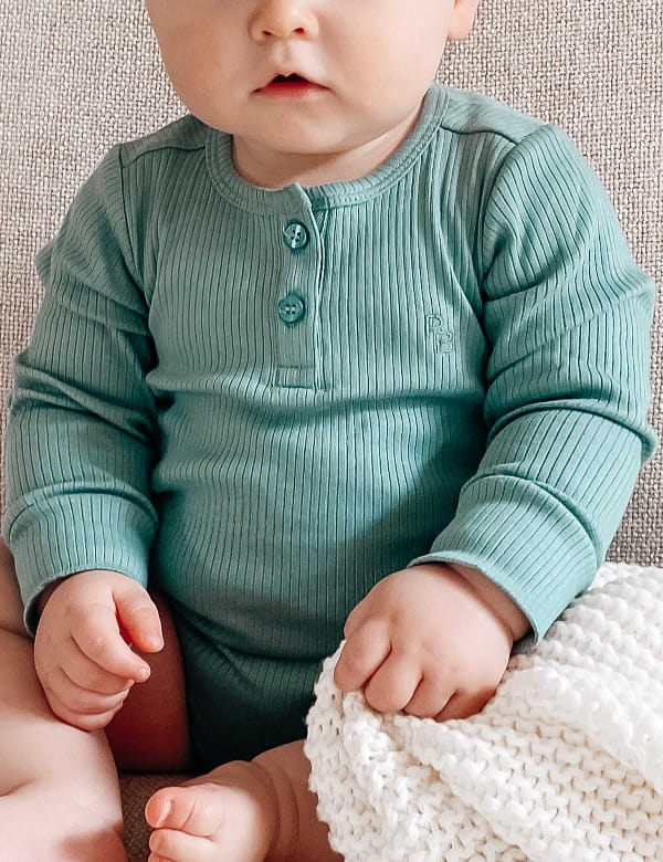 Baby wearing a teal ribbed onesie sitting on a textured surface with a white blanket.