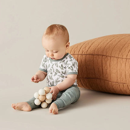 Baby sitting on a beige floor with a wooden toy, wearing a white shirt with green patterns by Sapling Child