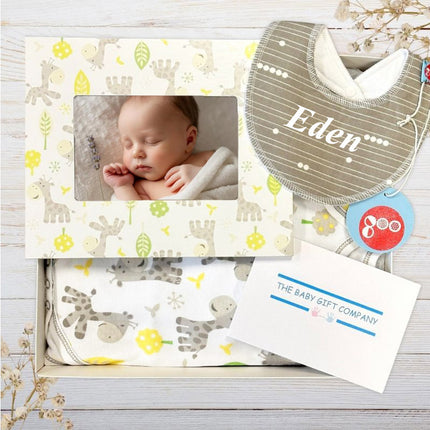 Baby gift set with grey personalised baby bib, photo frame, and blanket featuring animal patterns on a wooden surface.