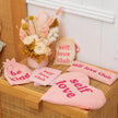 Pink 'self love club' items including bags, cards, and a heart-shaped pillow on a wooden surface.