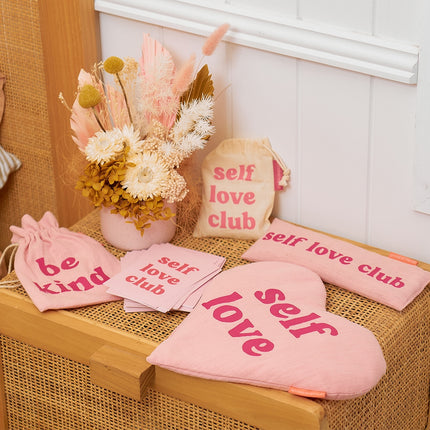 Pink 'self love club' items including bags, cards, and a heart-shaped pillow on a wooden surface.