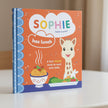 Book cover of 'Sophie la girafe' with a giraffe character and food items on an orange and blue background.