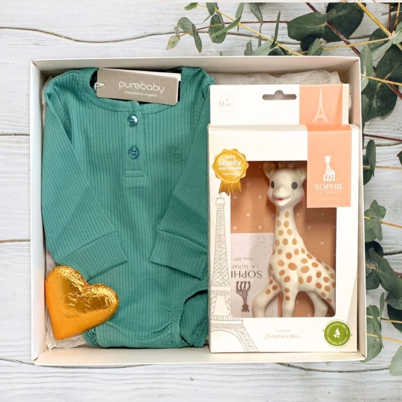 Baby gift set with teal cardigan, gold heart chocolate, and Sophie the Giraffe toy in gift box packaging on a wooden surface.