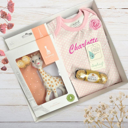 Baby gift set with sophie giraffe toy, pink top, rose , chocolates in a box on a wooden surface
