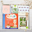 Collection of baby products including a book, sophie giraffe teether, and other items in a box with a white background and greenery.