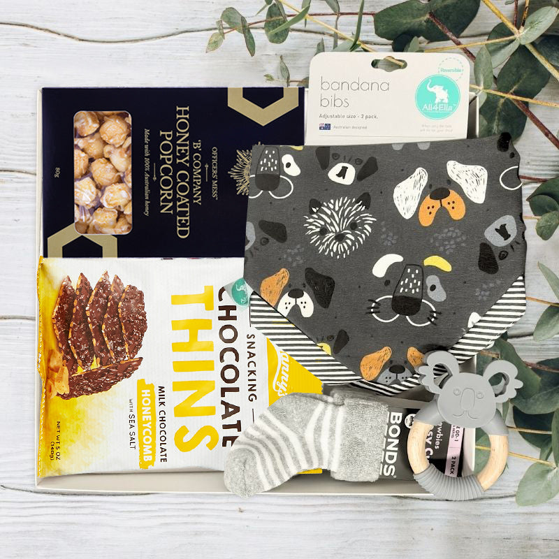 Gift box with popcorn, chocolate, bandana bib, socks, and teether on a wooden surface with greenery.