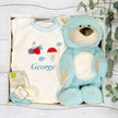 Baby gift set with blue teddy bear, white shirt with graphics, and socks in a box on a wooden surface.
