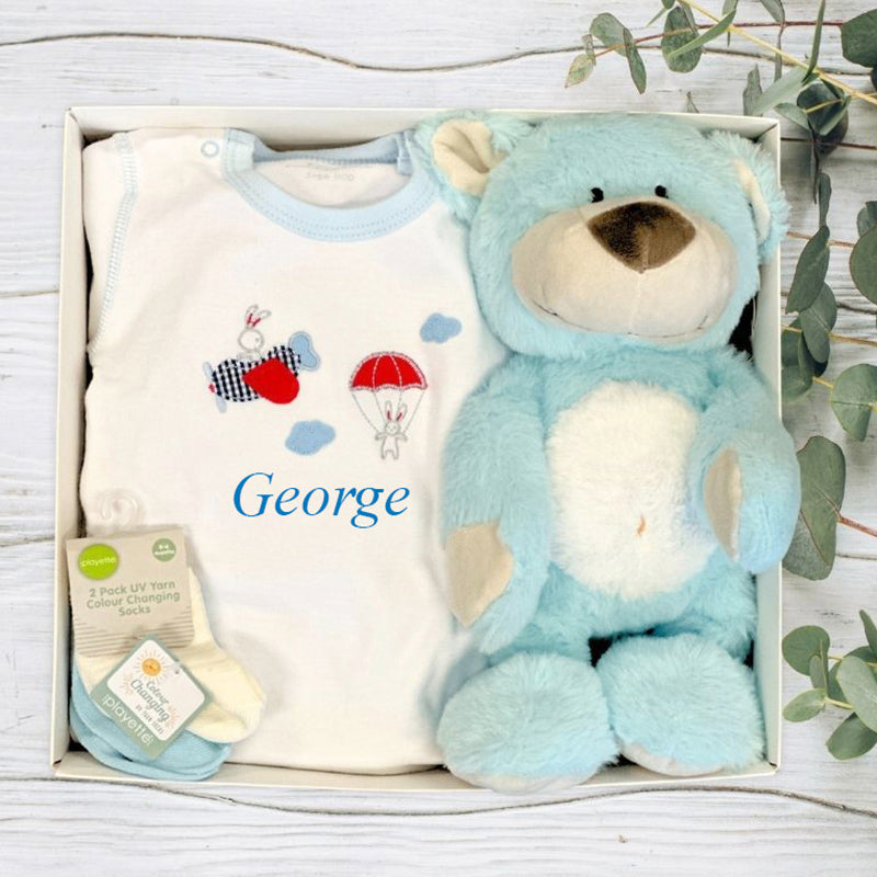 Baby gift set with blue teddy bear, white shirt with graphics, and socks in a box on a wooden surface.