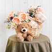 Wicker basket with flowers, teddy bear, and blanket on a green surface against a white wooden background