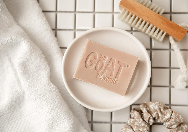 Bar of soap with 'The Goat Skincare' branding on a white plate, surrounded by a towel and brush on a tiled surface.